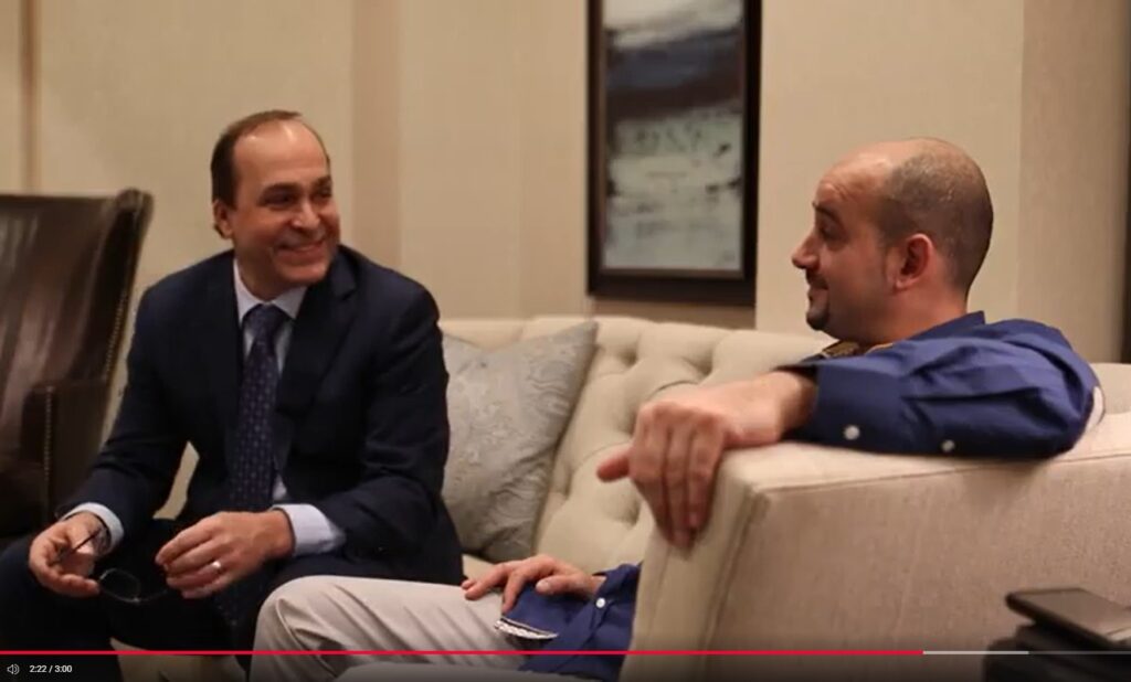 Two men sitting on a couch, engaged in a friendly conversation, wearing business attire in a cozy setting.