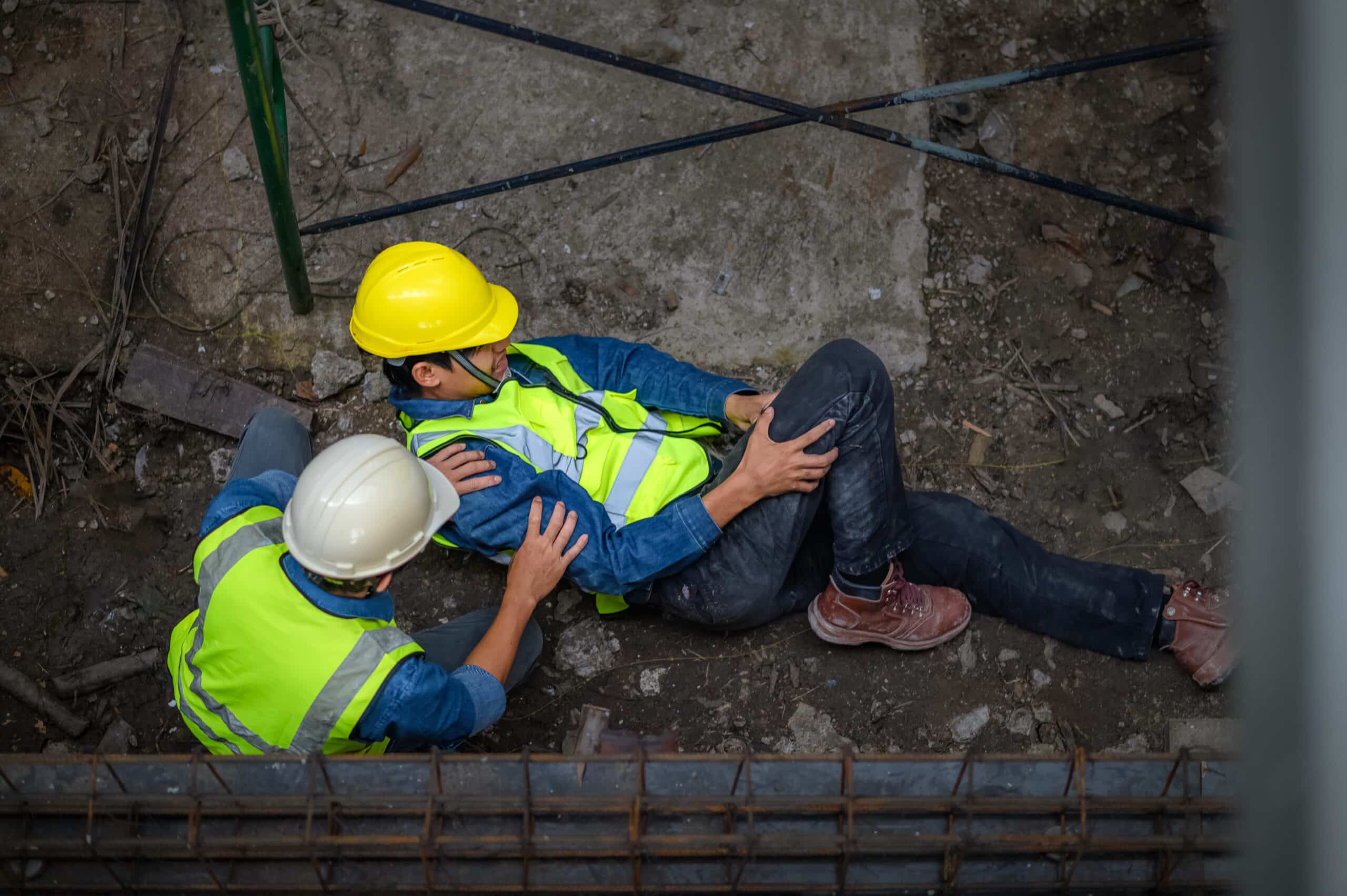 Two construction workers in safety gear assist an injured colleague on a construction site, highlighting workplace safety.